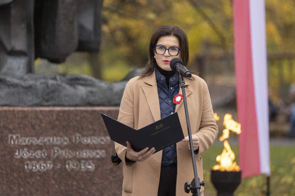 Kobieta z kotylionem w barwach narodowych wygłasza przemówienie przy mikrofonie podczas uroczystości przed pomnikiem Józefa Piłsudskiego. W tle widoczne są elementy pomnika, płomień symbolicznego znicza oraz biało-czerwone dekoracje.
