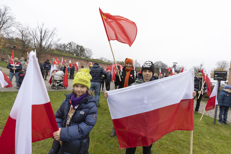 Dzieci i dorośli z biało-czerwonymi flagami uczestniczą w patriotycznym spotkaniu na Górze Gradowej. W tle widać budynki forteczne oraz liczne grupy osób z chorągiewkami i symbolami narodowymi.
