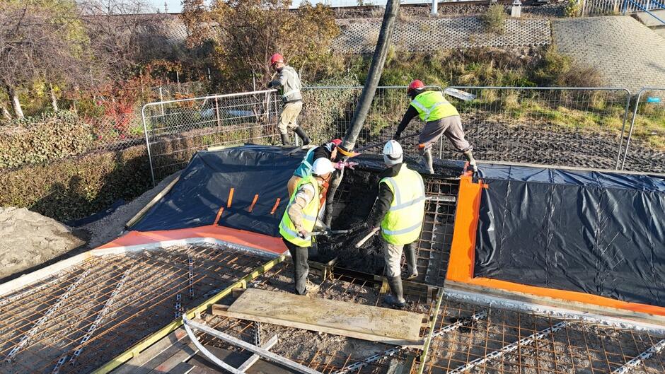 na zdjęciu robotnicy w jaskrawych strojach pracowniczych, część betonuje, część się przygląda