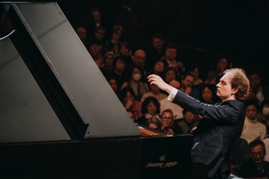 Pianista przy fortepianie, w trakcie koncertu. W tle jest widownia wypełniona publicznością