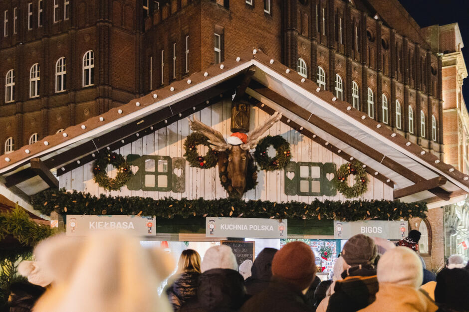 Tłum ludzi gromadzi się przed świątecznym stoiskiem z tradycyjną kuchnią polską, ozdobionym girlandami i wieńcami bożonarodzeniowymi. Nad wejściem wisi głowa łosia w czerwonej czapce Mikołaja, dodając stoisku humorystycznego, zimowego charakteru.
