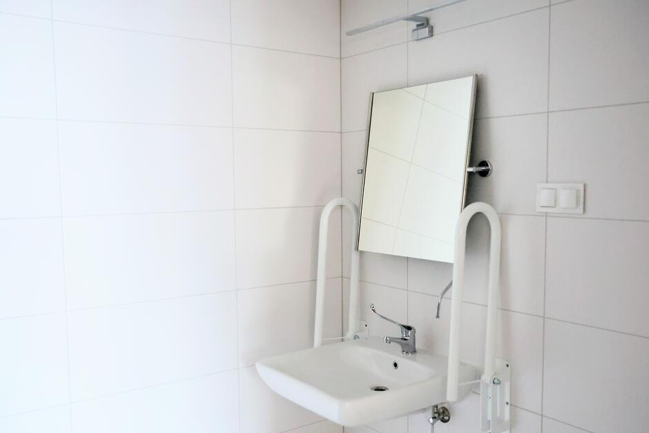 na zdjęciu łazienka z białymi kaflami na ścianach, widać też wiszące lustro i poręcze dla niepełnosprawnych