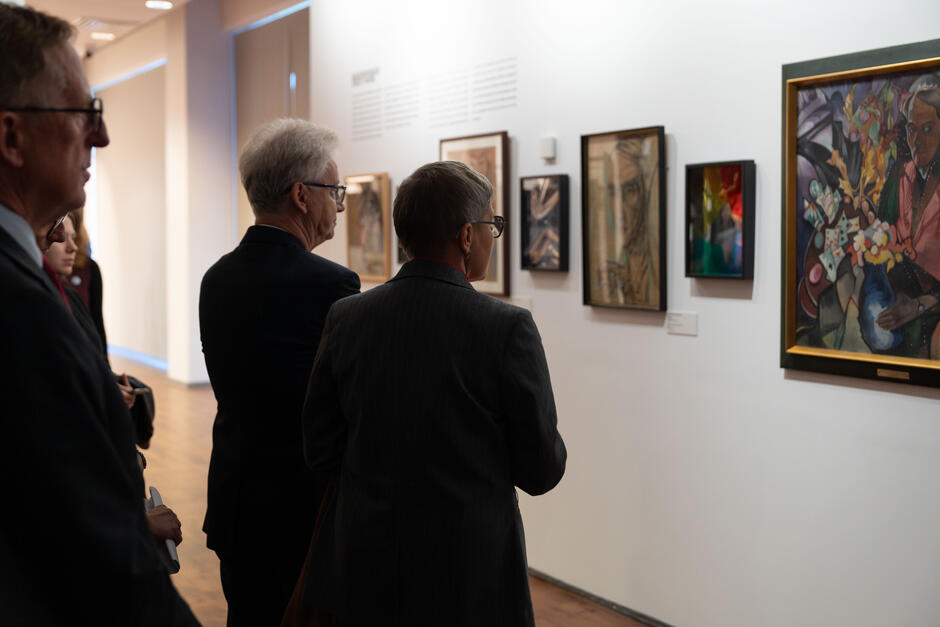 Zwiedzający w eleganckich strojach oglądają obrazy zawieszone na jasnej ścianie galerii. W skupieniu analizują szczegóły ekspresyjnych kompozycji i różnorodnych technik artystycznych.
