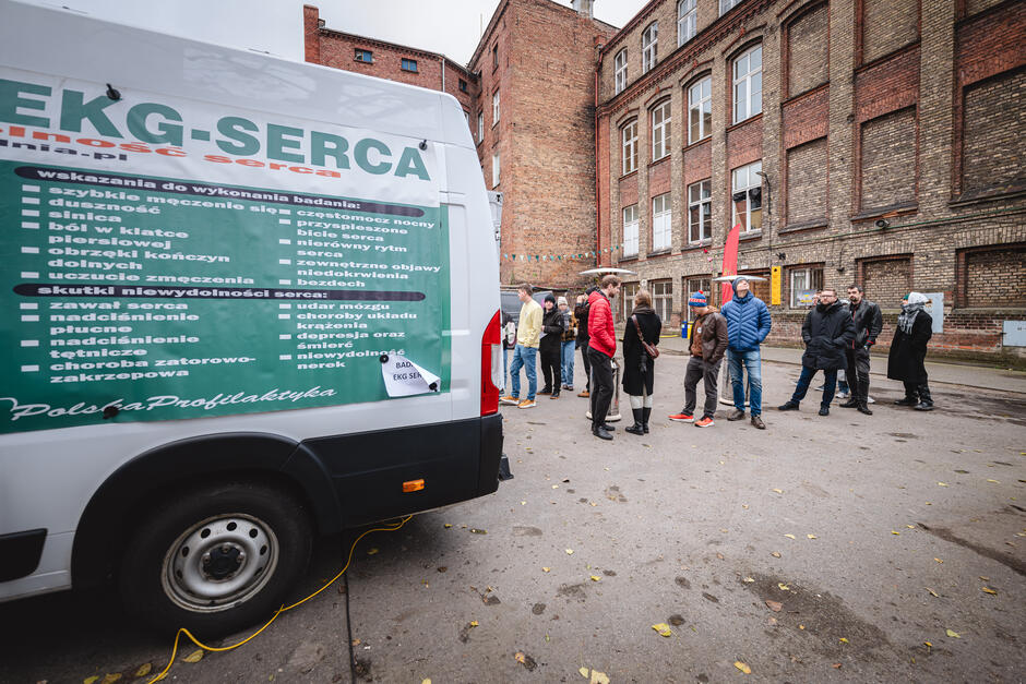 Grupa osób stoi w kolejce przed mobilnym punktem badań EKG serca zaparkowanym na podwórzu między budynkami podczas akcji profilaktycznej.