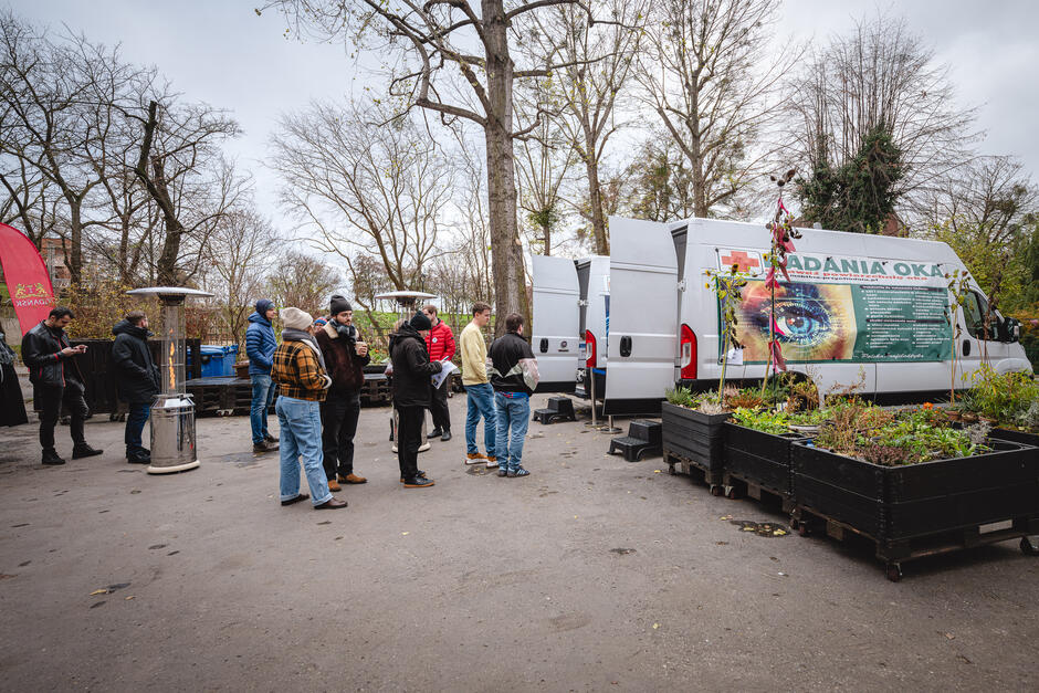 Grupa osób stoi w kolejce przed mobilnym punktem badań oczu zaparkowanym na podwórzu koło donic z kwiatami  podczas akcji profilaktycznej.