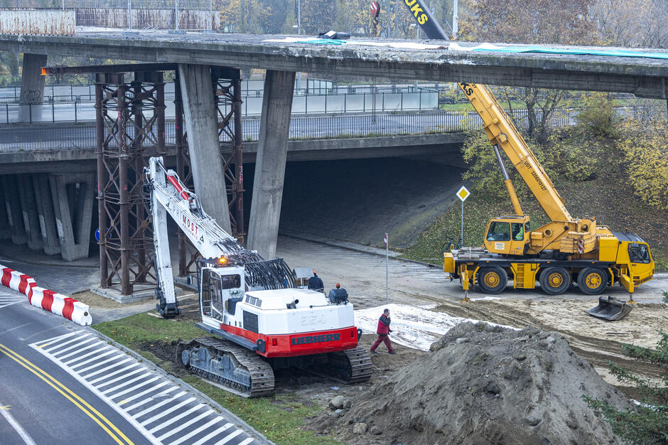 Na zdjęciu widać prace rozbiórkowe przy betonowej kładce, gdzie ciężki sprzęt – koparka gąsienicowa i żuraw – przygotowują konstrukcję do demontażu. Robotnicy działają u podstawy obiektu, a teren zabezpieczono barierami i podporami podtrzymującymi część przeprawy. 