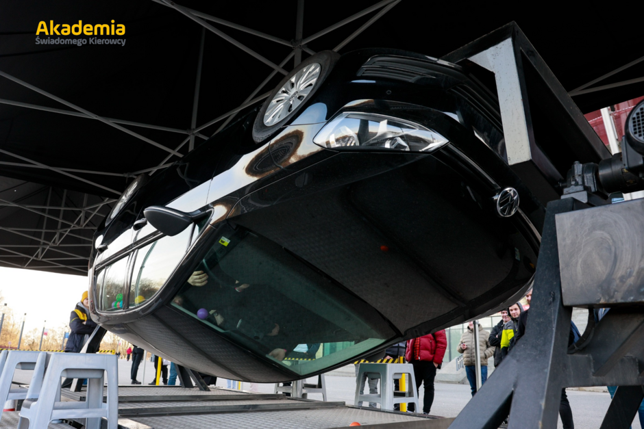 Symulator dachowania z czarnym samochodem osobowym ustawionym do góry kołami, używany w ramach Akademii Świadomego Kierowcy. W tle widoczne osoby obserwujące pokaz