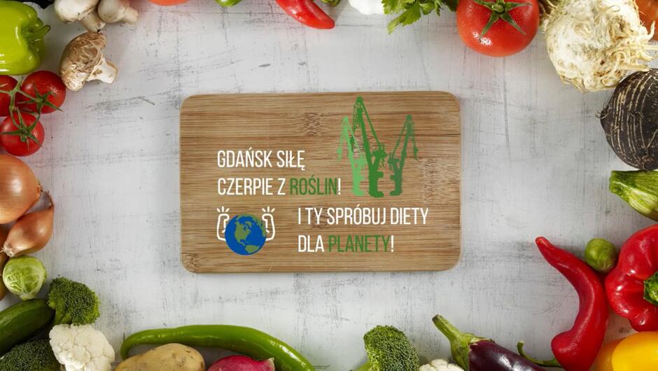 Na drewnianej desce widnieje hasło zachęcające do diety roślinnej, uzupełnione grafiką przedstawiającą żurawie stoczniowe oraz symbol Ziemi. Wokół deski ułożono różnorodne warzywa, tworząc kolorową i naturalną kompozycję.
