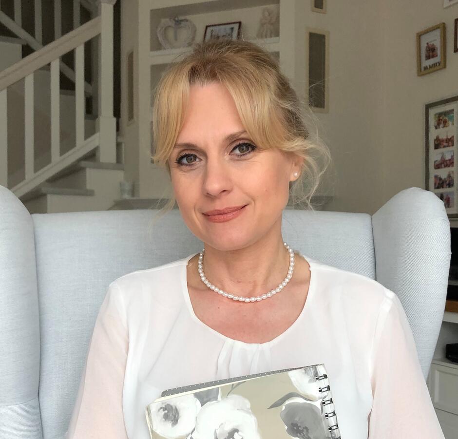 kobieta siedząca na jasnym fotelu, ubrana w elegancką bluzkę, trzymająca notes w kwiatowy wzór; w tle wnętrze z dekoracjami i schodami.