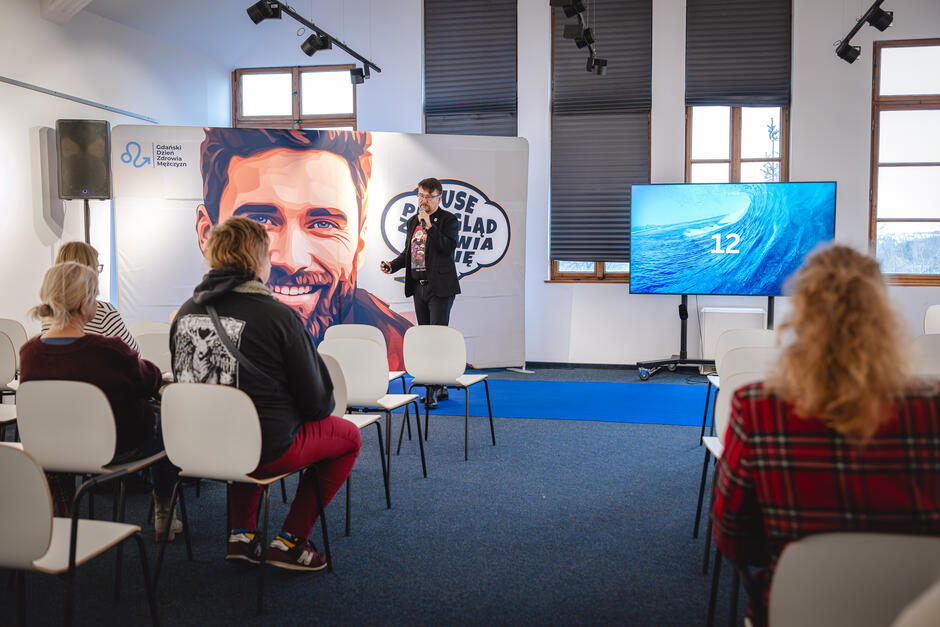 Prezentacja w sali konferencyjnej z uczestnikami siedzącymi na białych krzesłach, prowadzący stoi przy ściance z grafiką i napisem, obok duży ekran wyświetlający obraz fali