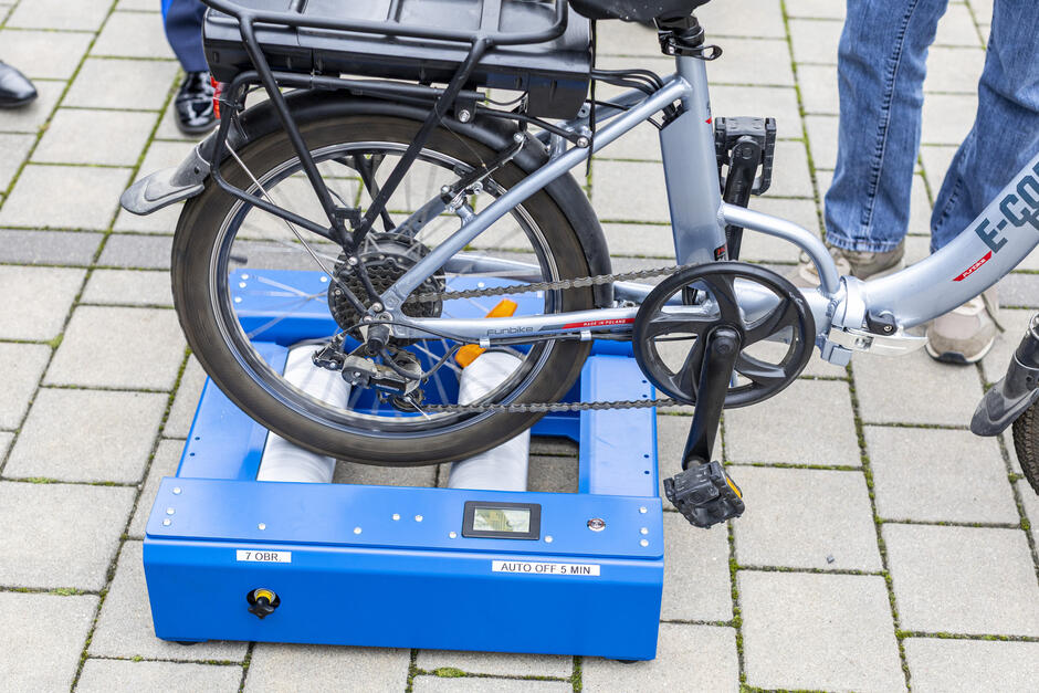 na zdjęciu koło roweru ustawione na urządzeniu w kolorze niebieskim, leży ono na chodniku