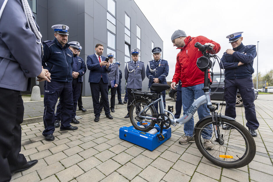 na zdjęciu stoi kilku umundurowanych policjantów, stoją w półokręgu, przyglądają się mężczyźnie w czerwonej kurtce który prezentuje urządzenie do mierzenia roweru