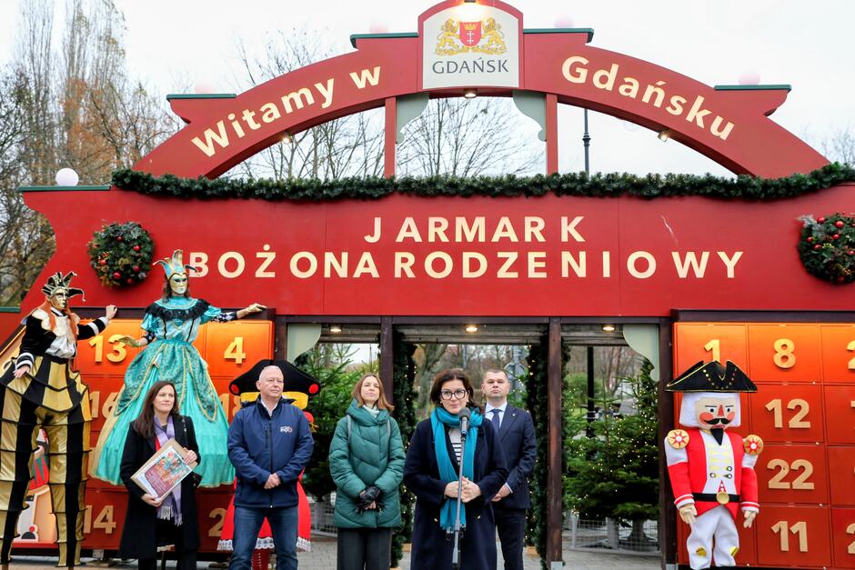 na zdjęciu sześc osóbstoi w rzędzie, w zimowych strojach, za nimi kolorowa, jarmarkowa brama adwentowa