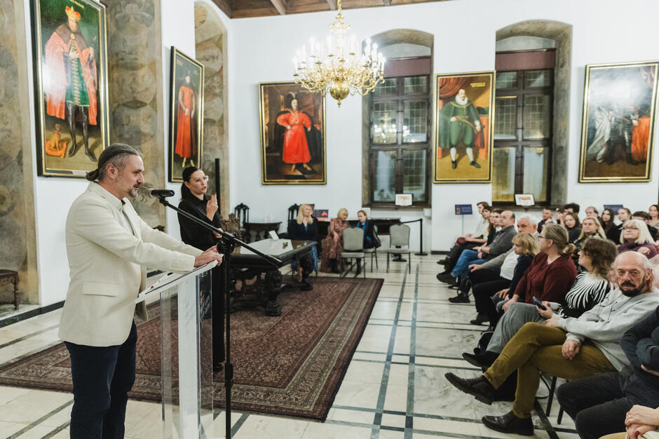 W dużej, reprezentacyjnej sali z wystrojem i obrazami sprzed wieków ludzie siedzą na krzesłach i słuchają mężczyzny, który stoi przed nimi na mównicy.