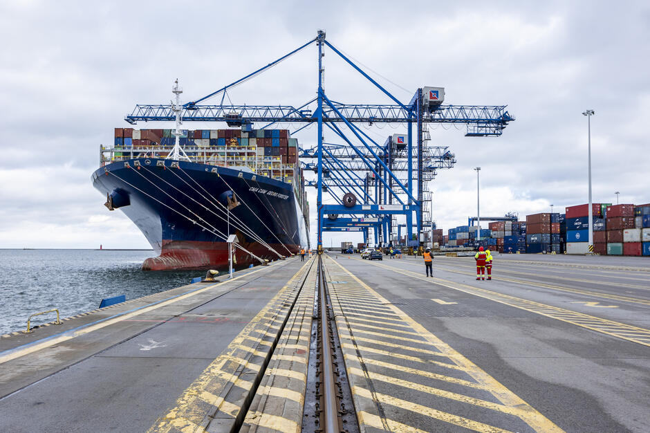 Duży statek zacumowany przy betonowym nabrzeżu, nad nim pracują suwnice, które wyładowują kontenery. Na pierwszym planie widoczne tory
