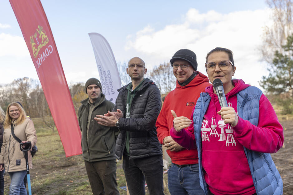 na zdjęciu kobieta mówi do mikrofonu, obok niej stoją w rzędzie trzej mężczyźni, wszyscy w średnim wieku