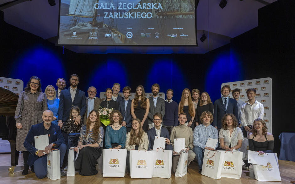 Na zdjęciu widać dużą grupę uśmiechniętych uczestników i laureatów stojących oraz siedzących na scenie podczas uroczystej gali żeglarskiej, trzymających dyplomy i torby z herbem Gdańska. W tle widoczny jest ekran z napisem „Gala Żeglarska Zaruskiego” oraz logotypami partnerów wydarzenia. 