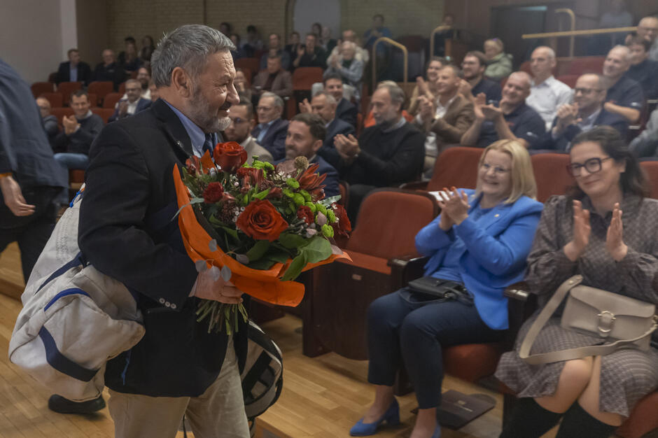 Na zdjęciu widać mężczyznę niosącego duży bukiet czerwonych kwiatów i torbę żeglarską, idącego wzdłuż rzędów krzeseł w sali pełnej widzów. Publiczność bije brawo i uśmiecha się, wyrażając uznanie i ciepłe przyjęcie. 
