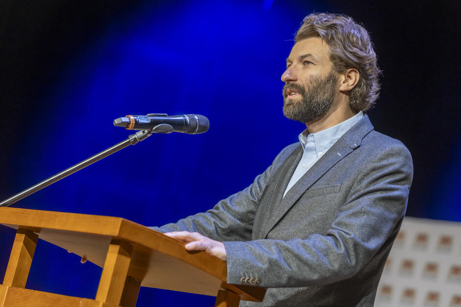 Na zdjęciu widać mężczyznę przemawiającego z mównicy, stojącego przy mikrofonie na oświetlonej, niebieskim światłem scenie. Ubrany w elegancką marynarkę i koszulę, mówi z zaangażowaniem, spoglądając w kierunku publiczności. 