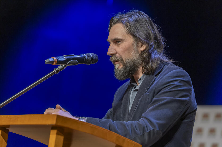 Na zdjęciu widać mężczyznę przemawiającego z mównicy, stojącego przy mikrofonie na oświetlonej, niebieskim światłem scenie. Ubrany w elegancką marynarkę i koszulę, mówi z zaangażowaniem, spoglądając w kierunku publiczności. 