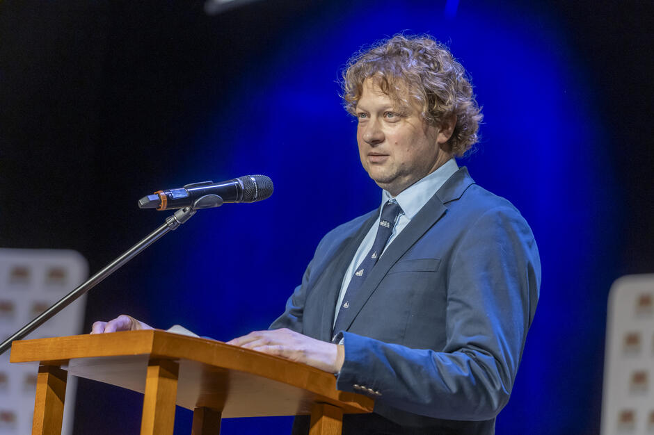 Na zdjęciu widać mężczyznę przemawiającego z mównicy, stojącego przy mikrofonie na oświetlonej, niebieskim światłem scenie. Ubrany w elegancką marynarkę i koszulę, mówi z zaangażowaniem, spoglądając w kierunku publiczności. 