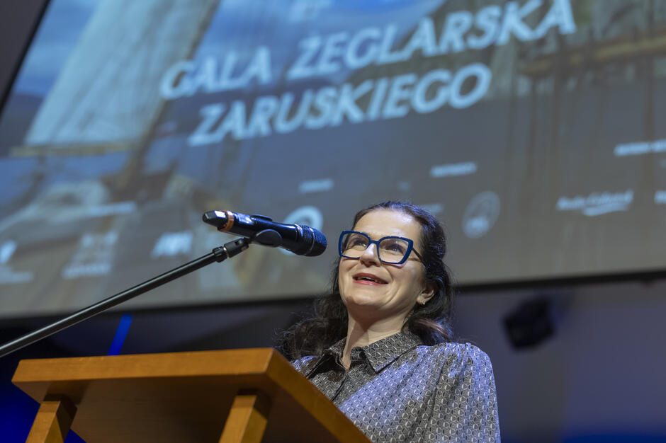 Na zdjęciu widać kobietę przemawiającą z mównicy, stojącą przy mikrofonie na tle dużego ekranu z napisem „Gala Żeglarska Zaruskiego”. Uśmiecha się i wygląda na zaangażowaną, a sceniczne światła podkreślają uroczysty charakter wydarzenia. 