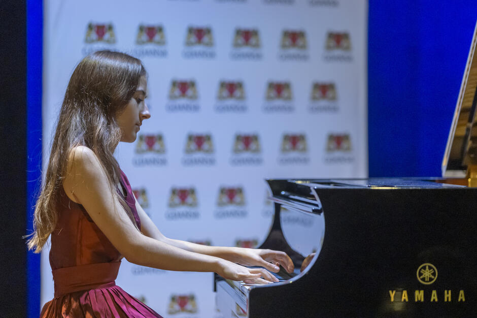 Na zdjęciu widać młodą pianistkę ubraną w elegancką bordową suknię, skupioną na grze podczas występu na fortepianie. Instrument znajduje się na scenie z tłem ozdobionym herbem Gdańska, co podkreśla uroczysty charakter wydarzenia. 