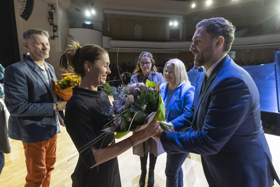 Na zdjęciu widać grupę osób na scenie podczas uroczystości, w chwili wręczania bukietów kwiatów. Uczestnicy uśmiechają się i gratulują sobie nawzajem, a atmosfera jest ciepła i pełna uznania. 