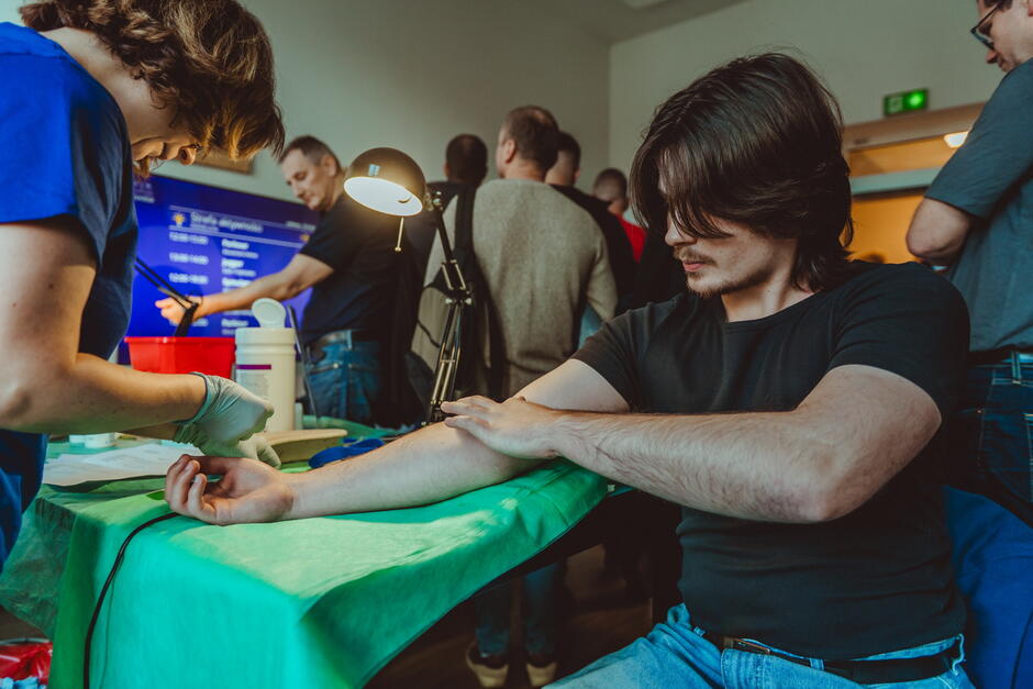 na zdjęciu młody mężczyzna, brunet, siedzi przy stole, wystawia lewa rękę do badania poboru krwi