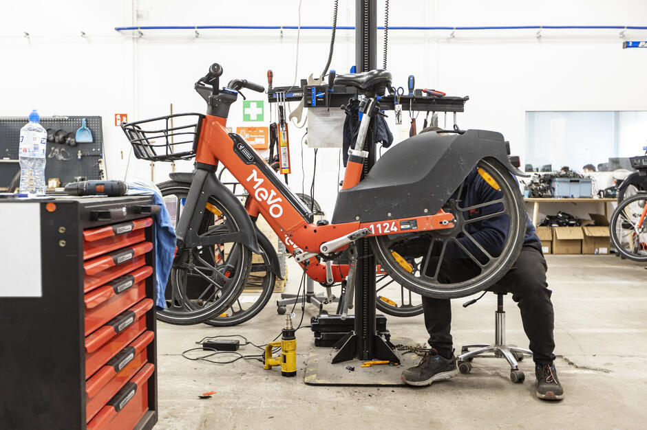 Na zdjęciu widać rower miejski Mevo umieszczony na stanowisku serwisowym w warsztacie, gdzie mechanik pracuje przy jego tylnym kole. Wokół widoczne są narzędzia, części i wyposażenie techniczne, podkreślające proces konserwacji i naprawy sprzętu. 