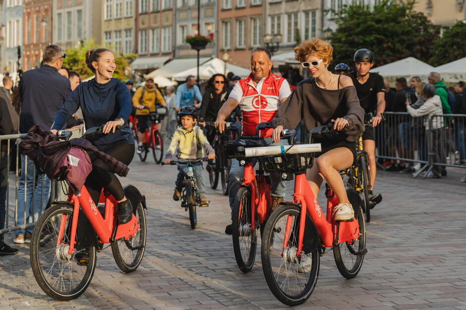 Na zdjęciu widać grupę uśmiechniętych osób jadących na czerwonych rowerach miejskich Mevo ulicą w centrum miasta, w otoczeniu zabytkowych kamienic i licznych przechodniów. Atmosfera jest radosna i pełna energii, a uczestnicy sprawiają wrażenie, że wspólna przejażdżka sprawia im dużo przyjemności. 