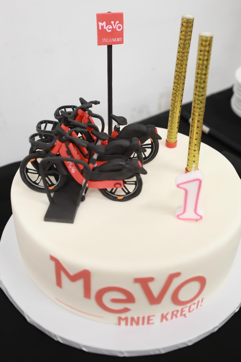 Na zdjęciu widać okrągły tort ozdobiony miniaturowymi modelami rowerów Mevo ustawionymi na wierzchu niczym w stacji dokującej oraz świeczką w kształcie cyfry 1. Dekoracje sugerują świętowanie pierwszej rocznicy systemu, a całość utrzymana jest w charakterystycznej biało-czerwonej kolorystyce marki. 