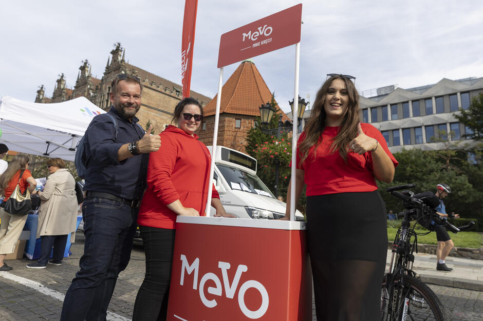 Na zdjęciu widać trzy uśmiechnięte osoby stojące przy czerwonym stoisku promocyjnym Mevo, pokazujące gest kciuka w górę podczas wydarzenia plenerowego w centrum miasta. W tle znajdują się zabytkowe budynki, namioty wystawowe oraz uczestnicy imprezy, tworząc radosną atmosferę. 