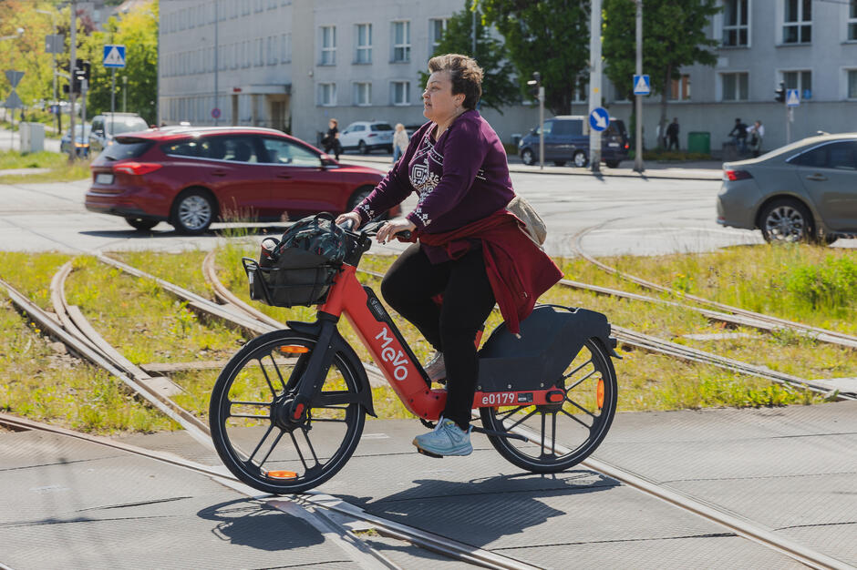 Na zdjęciu widać kobietę jadącą na czerwonym rowerze miejskim Mevo, przejeżdżającą przez tory tramwajowe na ruchliwej miejskiej ulicy. W tle widać samochody, budynki oraz sygnalizację świetlną, co podkreśla codzienny charakter przejazdu.
