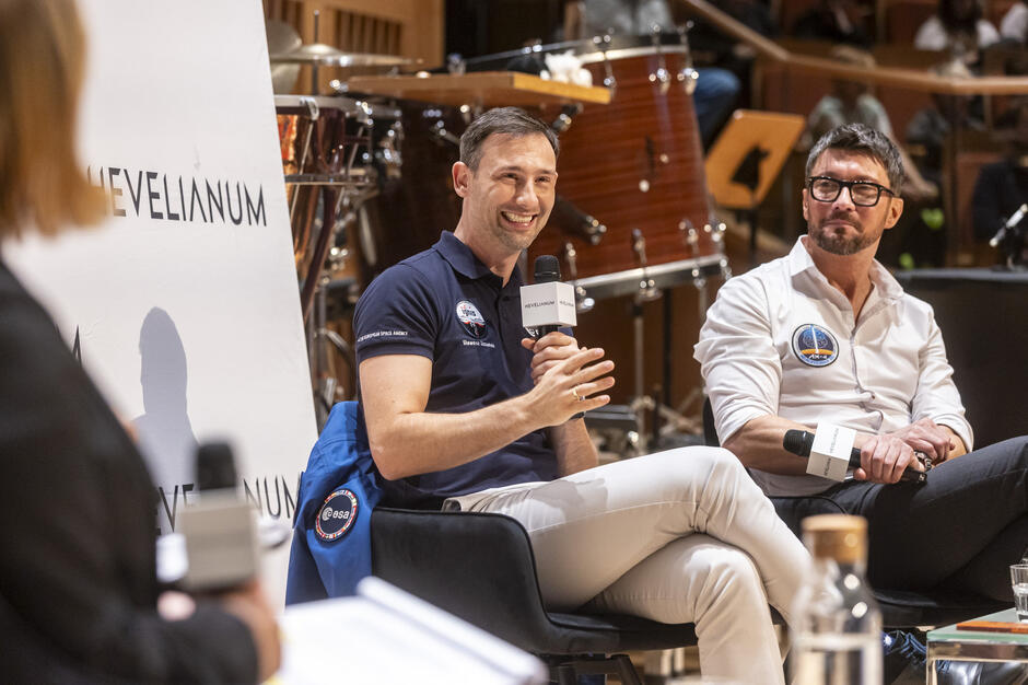 Na zdjęciu widać dwóch mężczyzn siedzących na scenie podczas spotkania lub panelu dyskusyjnego, trzymających mikrofony i rozmawiających z publicznością. Jeden z nich uśmiecha się i gestykuluje w trakcie wypowiedzi, a w tle znajdują się instrumenty perkusyjne, co sugeruje, że wydarzenie odbywa się w sali koncertowej lub audytoryjnej.
