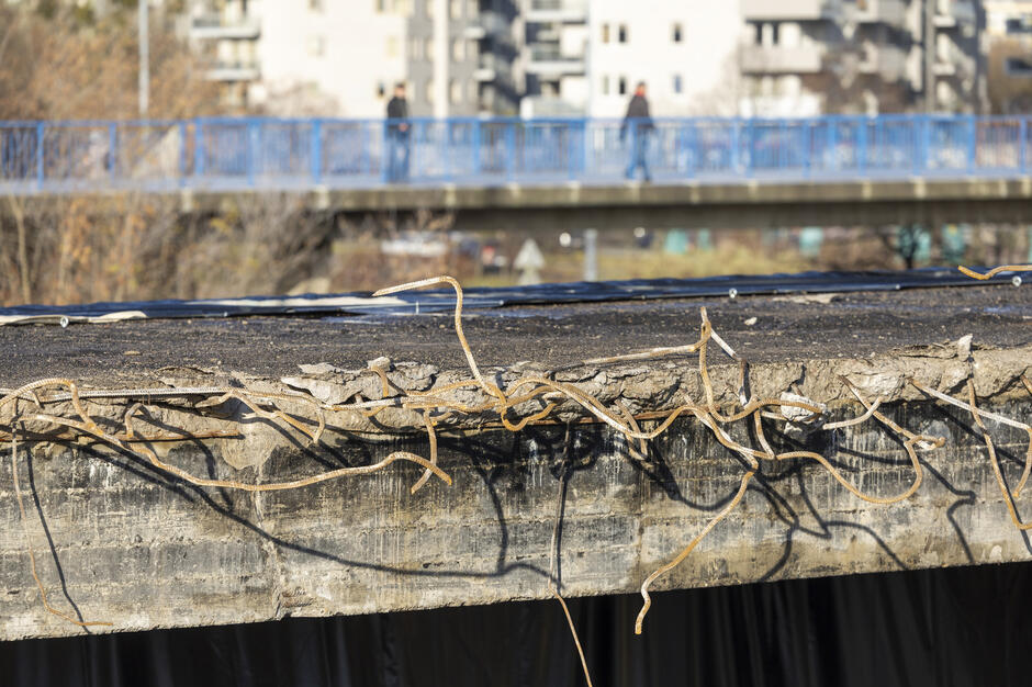 Na zdjęciu widać fragment zniszczonej betonowej konstrukcji z wystającymi, poskręcanymi prętami zbrojeniowymi. W tle znajduje się niebieska kładka dla pieszych oraz zabudowa mieszkaniowa, lekko rozmyta.
