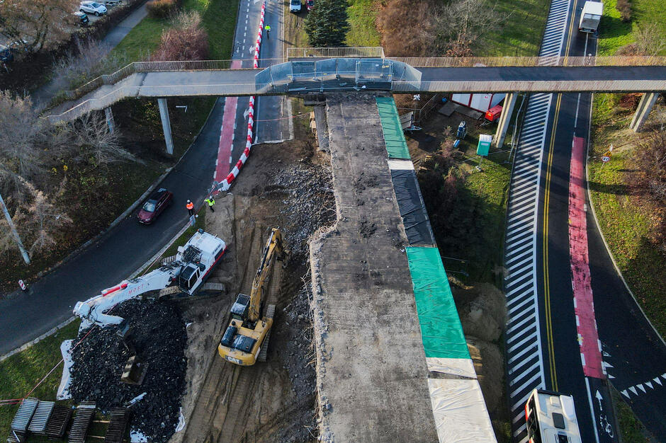 Zdjęcie zrobione z góry pokazuje rozbiórkę betonowej estakady, przy której pracują koparki oraz ekipa budowlana. Obok widać czynny wiadukt dla pieszych oraz przejeżdżające samochody na biegnących pod nim jezdniach.

