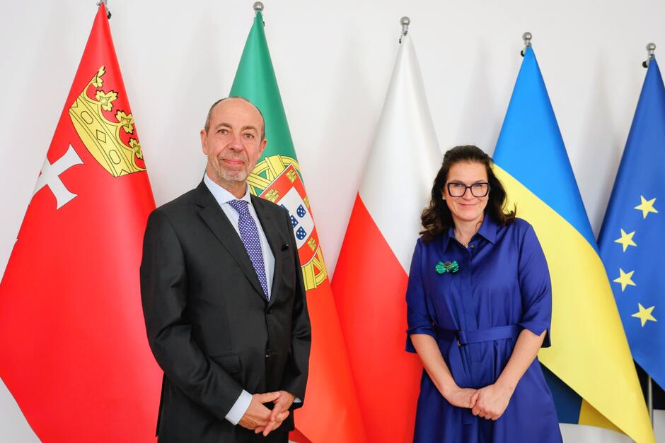 Mężczyzna po lewej, kobieta po prawej. Stoją na tle flag Polski i Portugalii, a także Gdańska i Unii Europejskiej 