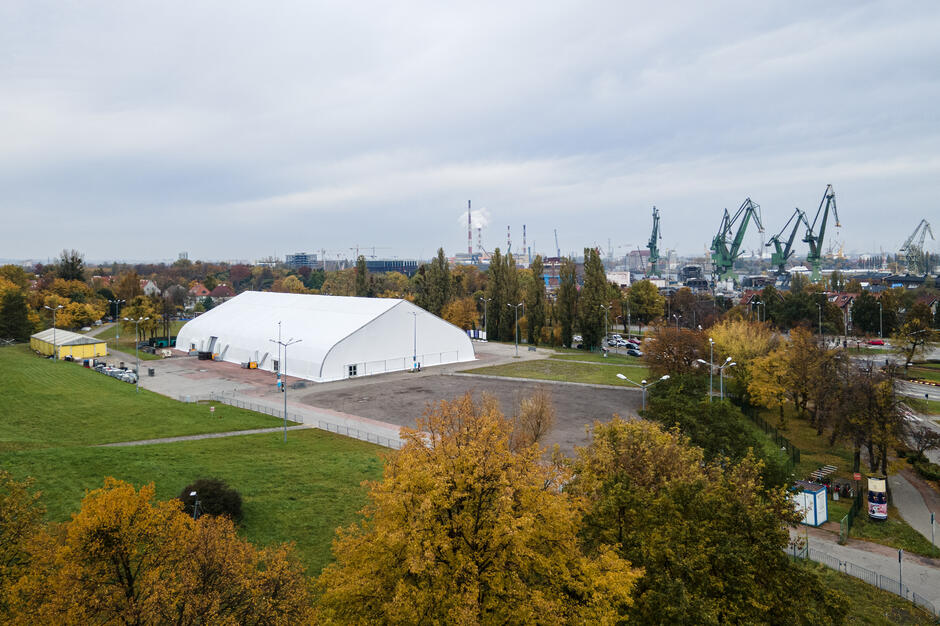 Na zdjęciu widoczny jest duży biały namiot ustawiony na rozległym placu otoczonym zielenią i jesiennymi drzewami. W tle rozciąga się panorama miasta z charakterystycznymi żurawiami stoczniowymi oraz elementami przemysłowej infrastruktury.

