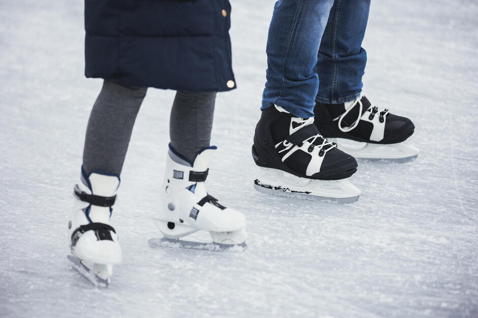 Na zdjęciu widać dwie osoby stojące na lodzie, ujęte w kadrze od nóg w dół, w łyżwach rekreacyjnych. Jedna z nich ma biały model, druga czarno-biały, a powierzchnia lodowiska jest lekko porysowana od jazdy.
