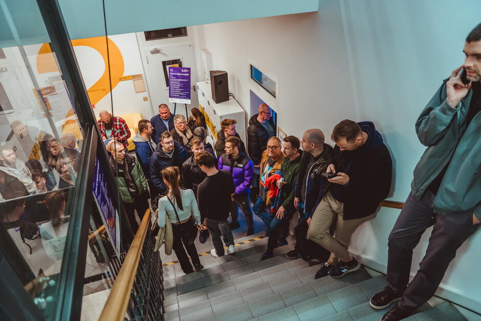 Grupa osób stoi w kolejce na klatce schodowej w nowoczesnym budynku. Widoczne są szare schody, metalowa poręcz oraz szklana balustrada po lewej stronie. W tle znajduje się biała ściana z dużą pomarańczową cyfrą „2” oraz fioletowa tablica informacyjna. Jedna osoba opiera się o ścianę po prawej stronie i rozmawia przez telefon