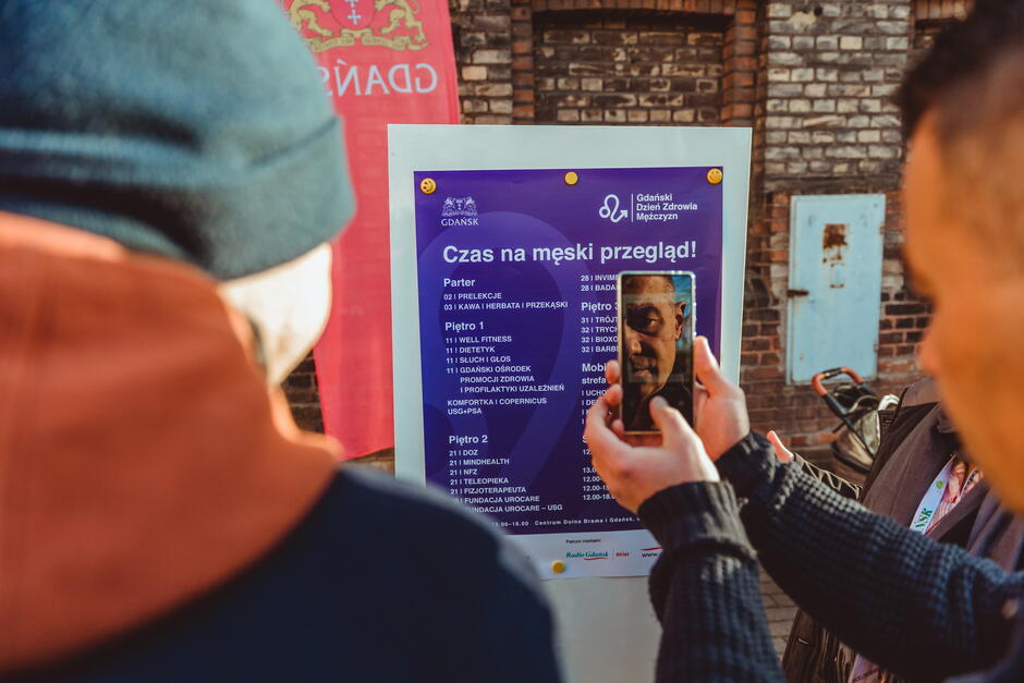 Dwie osoby stoją przed tablicą informacyjną z fioletowym plakatem zatytułowanym „Czas na męski przegląd!” dotyczącym Gdańskiego Dnia Zdrowia Mężczyzn. Na plakacie widoczne są szczegóły wydarzenia, w tym lokalizacja i harmonogram na różnych piętrach. Jedna z osób trzyma smartfon, na którego ekranie widać zdjęcie twarzy. W tle znajduje się ceglana ściana budynku oraz niebieskie drzwi