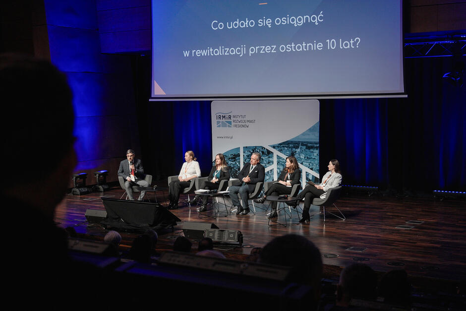 Kilka osób siedzi na fotelach na scenie i rozmawia, słucha ich publiczność.