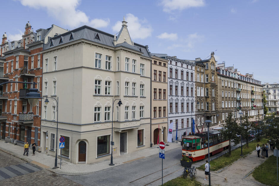  Scena uliczna w europejskim mieście. Na ulicy znajduje się kilka budynków, w tym duży biały budynek z czarnym dachem i wieżą zegarową na górze. Budynki są różnych rozmiarów i kolorów, z których niektóre są czerwone, białe i beżowe. Po prawej stronie jest czerwono-biały wózek na brukowanej ulicy. Ludzie mogą spacerować po chodniku i chodzić. W tle niebo jest niebieskie, widoczne są drzewa i inne budynki.