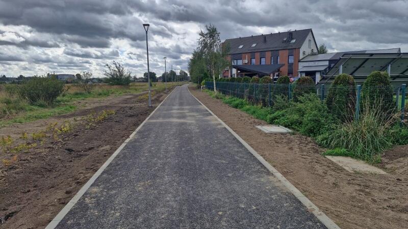 na zdjęciu droga rowerowa o czarnej nawierzchni, w tle budynek jednorodzinny