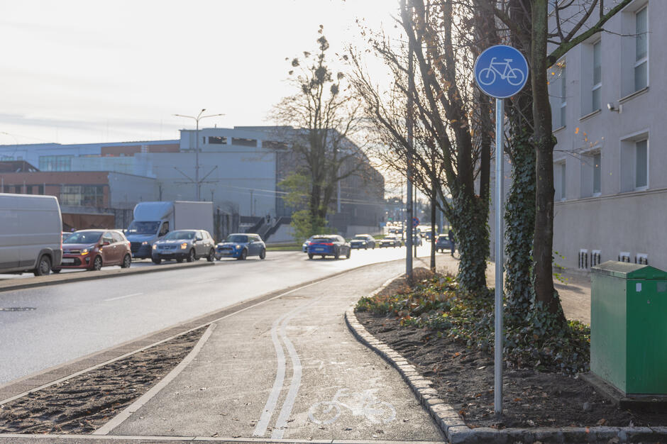 na zdjęciu droga rowerowa z białymi piktogramami przedstawiającymi rowery, o czarnej nawierzchni, po prawej widać niebieski znak drogowy, w tle po prawej jasny budynek, po lewej kilka samochodów na jezdni