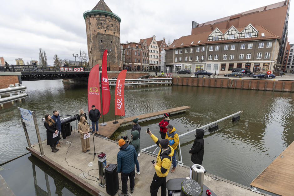W marinie żeglarskiej w mieście stoją ludzie na kei podczas konferencji prasowej.