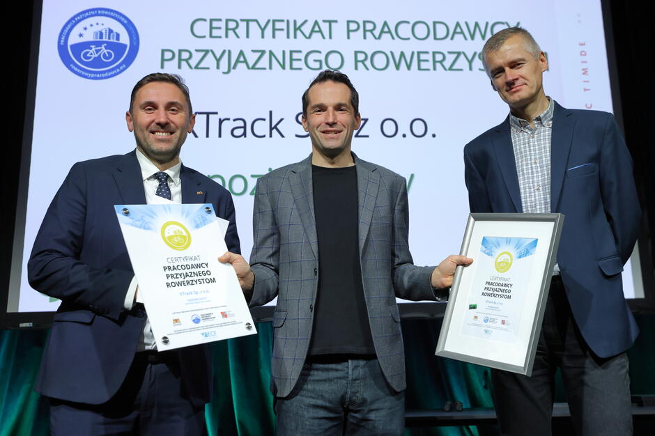 Na zdjęciu trzech mężczyzn stoi na scenie, trzymając w rękach certyfikaty „Pracodawcy Przyjaznego Rowerzystom”. W tle widać ekran z nazwą programu oraz logotypem inicjatywy rowerowej. 