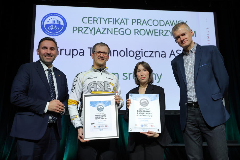 Na zdjęciu znajdują się cztery osoby stojące na scenie, prezentujące certyfikaty „Pracodawcy Przyjaznego Rowerzystom”. Za nimi widoczny jest ekran z logo programu i nazwą nagrodzonej organizacji.
