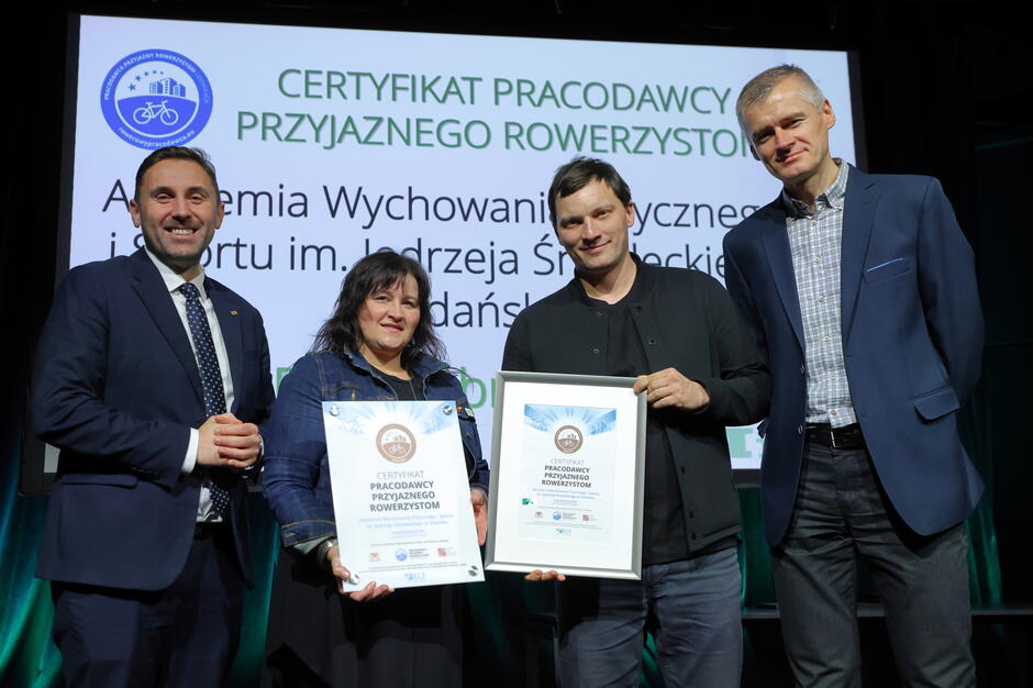 Na zdjęciu cztery osoby stoją na scenie, trzymając w dłoniach certyfikaty „Pracodawcy Przyjaznego Rowerzystom”. W tle znajduje się ekran z nazwą nagrodzonej instytucji oraz logo programu.
